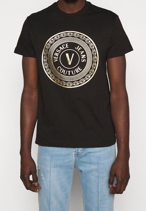 Schwarzes Baumwoll-T-Shirt mit einem zentralen goldenen runden Logo mit der Aufschrift "Versace Jeans Couture" und einem gemusterten Rand. Kurze Ärmel, Rundhalsausschnitt.