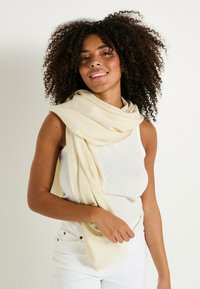 Bufanda tejida beige con una textura suave, caída de manera casual sobre los hombros, complementando una blusa blanca sin mangas y pantalones claros.