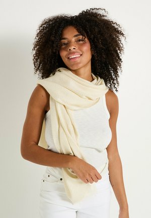 Écharpe tissée beige avec une texture douce, drapée de manière décontractée autour des épaules, complétant un top sans manches blanc et un pantalon léger.