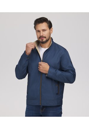 Mężczyzna o krótkich ciemnych włosach i brodzie zapinający niebieską kurtkę z wzorem na białą koszulę, stojący na tle jasnego, jednolitego tła.