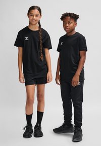 Dos niños llevan camisetas de manga corta y pantalones negros a juego. Las camisetas tienen un pequeño logotipo blanco. Ambos llevan calzado y calcetines negros.