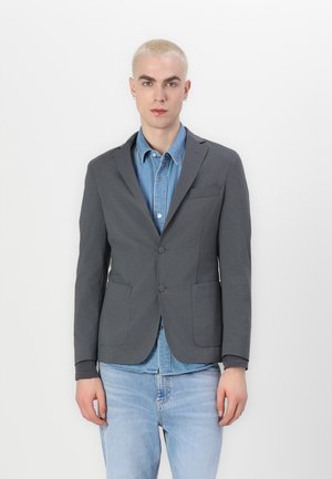 Blazer gris con un corte entallado, solapas en pico, dos bolsillos frontales y dos botones, llevado sobre una camisa de mezclilla de color azul claro y jeans claros.