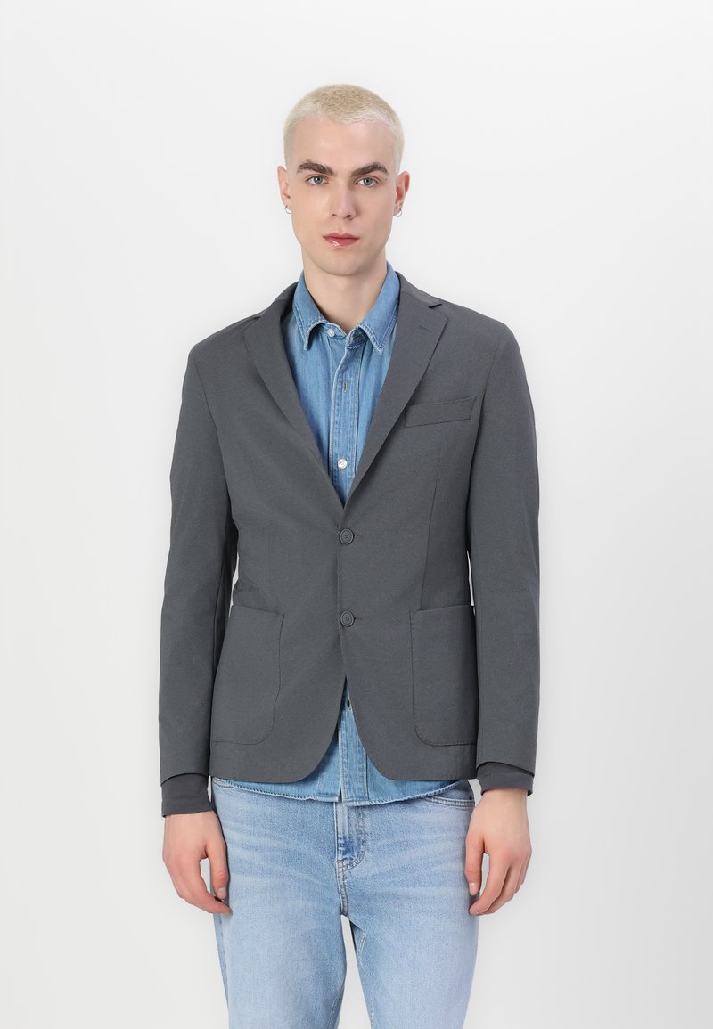 Grijze blazer met een tailored fit, notch-kraag, twee voorzakken en twee knopen, gedragen over een lichtblauw denimshirt en lichte jeans.