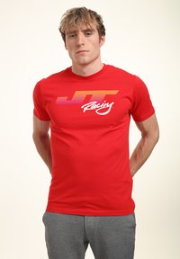 Červené tričko s krátkým rukávem a přiléhavým střihem. Obsahuje grafiku v barevném přechodu od oranžové po růžovou, která zobrazuje nápis "JT Racing" v bílém kurzívě.