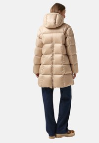 Beige Pufferjacke mit Kapuze, mit gesteppter Optik, Reißverschlussverschluss und Rückenstickerei. Kombiniert mit dunklen Jeans und braunen Stiefeln.