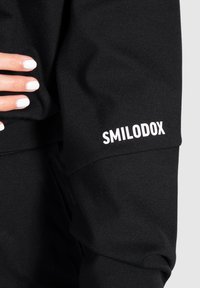 Zwarte sweatshirt met een gladde textuur. Heeft een wit "SMILODOX" logo op de mouw. Close-up van de stof en een hand die erop rust.