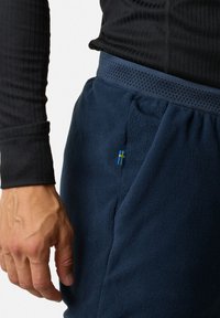 Marineblauwe fleece joggers met een gestructureerd oppervlak, elastische tailleband, zijzakken en een klein blauw-geel vlagdetail. Zwarte langekotige top zichtbaar.