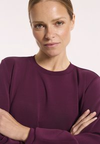 Bordowy longsleeve z gładką teksturą, okrągłym dekoltem i luźnym krojem, z subtelnymi przeszyciami na rękawach.