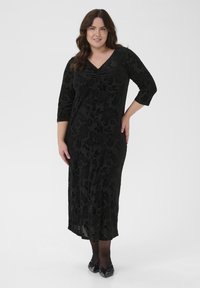 Fekete, térdig érő ruha virágos bársonyos textúrával, V-kivágással és háromnegyedes ujjakkal; fekete masnival díszített lapos cipővel párosítva.