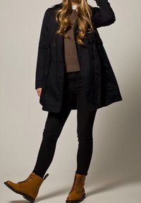 Czarny trenchcoat na brązowym swetrze i czarnych skinny jeansach. Beżowe, sznurowane botki za kostkę dodają kontrastu. Długie, falowane włosy dopełniają całość.