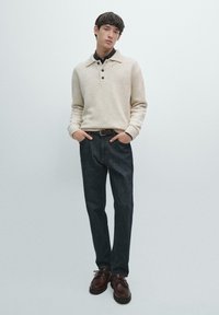 Beżowy wełniany sweter z kołnierzem, ciemne dżinsowe spodnie i brązowe skórzane buty. Strój charakteryzuje się luźnym krojem i subtelnymi fakturami.