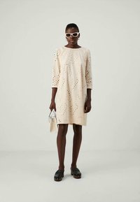 Ανοιχτό μπεζ φόρεμα με κοψίματα, τρία τέταρτα μανίκια και χαλαρή εφαρμογή. Συνδυασμένο με μαύρες ξύλινες τσάντες και λευκή τσάντα.