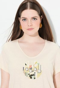 Beige T-shirt met korte mouwen, featuring ronde bloemenborduursels in diverse kleuren, met een wijd ronde hals en een zachte textuur.