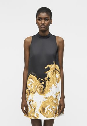 Mujer con vestido sin mangas en blanco y negro con patrones de remolinos barrocos dorados, de pie contra un fondo claro y liso.