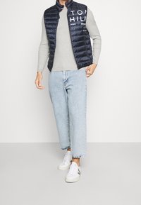 Gilet imbottito navy con dettaglio logo, maglione grigio a coste, jeans azzurri corti e scarpe da ginnastica bianche con dettagli grigi.