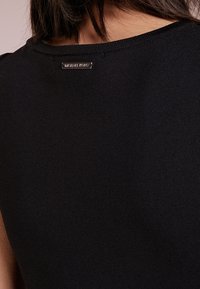 Haut noir à côtes avec une texture finement tissée et un petit logo métallique au niveau de l'encolure au dos. Design simple et ajusté.