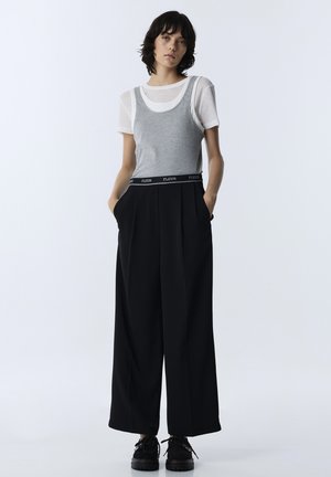 Giovane donna con capelli corti e scuri che indossa una t-shirt bianca sotto una canottiera grigia, pantaloni neri a gamba larga e scarpe nere con le mani nelle tasche.