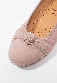 Chausson plat en daim rose clair avec un détail de nœud au niveau des orteils et un tissu froncé. L'intérieur présente une semelle perforée pour la respirabilité.