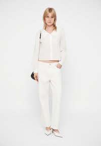 Camicia bianca corta con un grande colletto, chiusura frontale a bottoni, abbinata a pantaloni bianchi a gamba dritta e tacchi a punta, mentre tiene una borsa nera.