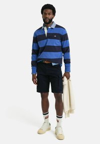 Camisa polo de manga larga a rayas en azul y navy, pantalones cortos cargo negros, zapatillas de crema y calcetines blancos con letras rojas. Atuendo casual con texturas superpuestas.