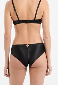 Set di lingerie nero con texture liscia in raso. Il reggiseno ha spalline regolabili e una chiusura a gancio. Le mutandine presentano un dettaglio decorativo con ritaglio sul retro.