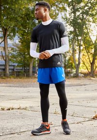 Maglietta sportiva nera a maniche corte, maniche lunghe bianche sotto, pantaloni corti blu, leggins neri e sneakers grigie con accenti arancioni.