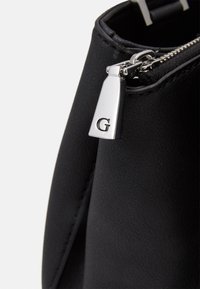 Mustat tekonahkalaukku, jossa on hopeanvärinen vetoketju ja metallinen riipus, jossa on kirjain "G".Teksturisoitu pinta, tyylikäs muoto ja minimalistisella designilla.