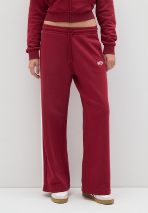 Pantaloni della tuta bordeaux in un fit rilassato, realizzati in tessuto morbido. Presentano una vita elasticizzata con coulisse e righe bianche sui lati.
