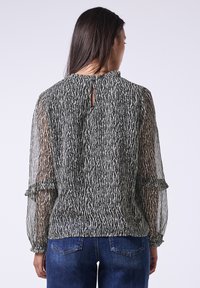 Schwarz-weiß gemusterte Bluse mit transparenten langen Ärmeln, Rüschensäumen und einem Schlüsselloch-Ausschnitt hinten, kombiniert mit dunkelblauen Jeans.