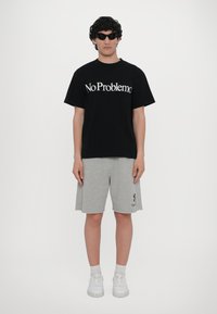 Svart bomull T-skjorte med hvit tekst "No Problemo", kombinert med grå bomullsshorts. Brukt med hvite sneakers og svarte solbriller.