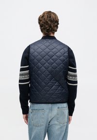 Άνδρας με καστανά σγουρά μαλλιά, φορώντας μια ναυτική γυμναστήριου γιλέκο πάνω από ένα μαύρο πουλόβερ με λευκές ρίγες στα μανίκια και ανοιχτό μπλε τζιν, στραμμένος μακριά.