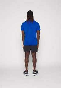 Blaues Sport-T-Shirt mit kurzen Ärmeln, kombiniert mit schwarzen Shorts. Getragen mit schwarzen und grauen Laufschuhen. Sicht von hinten.