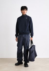 Felpa crewneck blu navy, pantaloni cargo e scarpe nere chunky. L'uomo tiene una borsa blu navy con manici neri, dotata di una tasca frontale. Texture liscia.