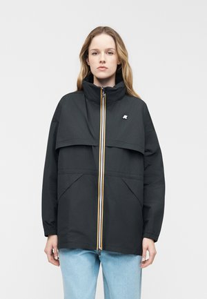 Jeune femme portant une veste noire zippée à col montant et un jean bleu clair, debout devant un fond uni clair.