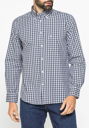 Uomo con camicia a maniche lunghe a quadri bianco e nero e jeans blu, che posa con una mano abbassata.