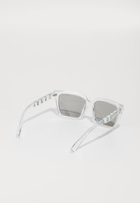 Ochelari de soare din plastic transparent cu lentile gri. Rame cu formă pătrată și brațe texturate, cu striații. Designul general este modern și minimalist.