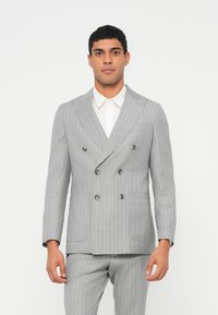 Giacca doppiopetto grigia a righe sottili con rever a incavo, due tasche frontali e bottoni grigi, abbinata a una camicia bianca con colletto.