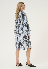 Obleka je izdelana iz lahkega materiala v svetlo modri barvi z črnimi abstraktnimi vzorci, ima dolge rokave in zategnjen pas, kombinirana s črnimi slingback petami.