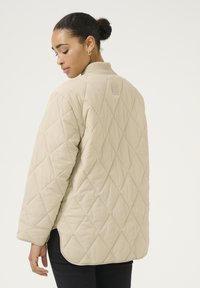 Beige gewatteerde jas met een geribbelde kraag, voorzien van een ruitpatroon en een logotag op de achterkant. Lange mouwen en een afgeronde zoom.