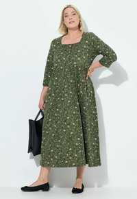 Abito maxi floreale verde con maniche a tre quarti, scollatura quadrata e vita arricciata. Abbinato a ballerine nere e una borsa tote.