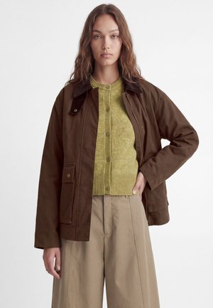 Femme aux longs cheveux bruns portant une veste marron, un pull vert boutonné et un pantalon beige à larges jambes, debout avec une main sur la hanche.