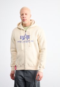 Béžová mikina na zips s fialovým logom "Alpha Industries". Má kapucňu so šnúrkou a predné vrecká, pričom sa vyznačuje hladkou, jemnou textúrou.