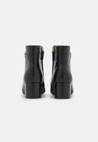 Botas de tobillo de cuero negro con tacón grueso, que cuentan con un zipper lateral y acentos metálicos dorados. Textura suave y diseño elegante.