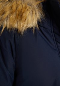Marineblå jakke med en faux fur-krave i lys brune. Stoffet ser glat ud med en shiny overflade; kraven har lange, bløde fibre.