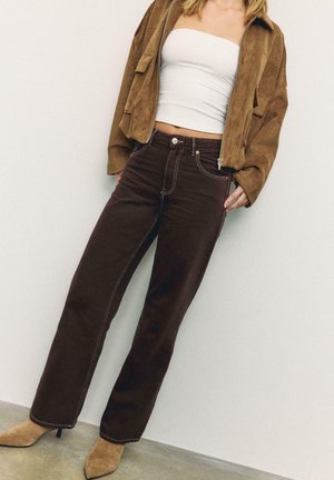 Donna che indossa una giacca in suede marrone, un top bianco senza spalline, jeans marrone scuro a vita alta e stivaletti color cognac con punta a punta, in piedi contro un muro bianco.