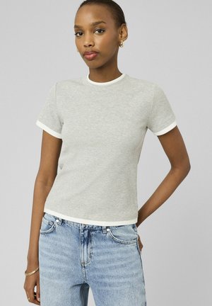 Jeune femme portant un haut gris clair côtelé à manches courtes avec un bord blanc et un jean taille haute bleu clair, posant devant un fond uni.