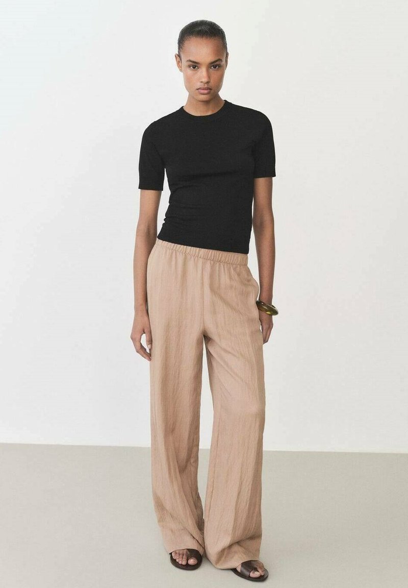 Czarny dopasowany top z krótkim rękawem w połączeniu z beżowymi spodniami typu wide-leg, wykonanymi z lekkiego materiału. Modelka nosi czarne sandały oraz prostą bransoletkę.