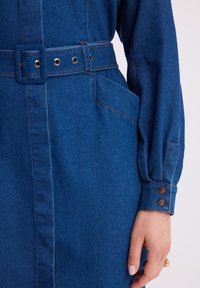 Dżinsowa sukienka z dopasowaną talią, niebieskiego materiału, z guzikami na mankietach i bocznymi kieszeniami. Posiada kontrastowe przeszycia oraz prostokątny pasek.