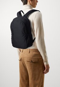 Zaino in tessuto nero con chiusura a zip e tasche laterali in rete. Texture liscia e spallacci ergonomici visibili.
