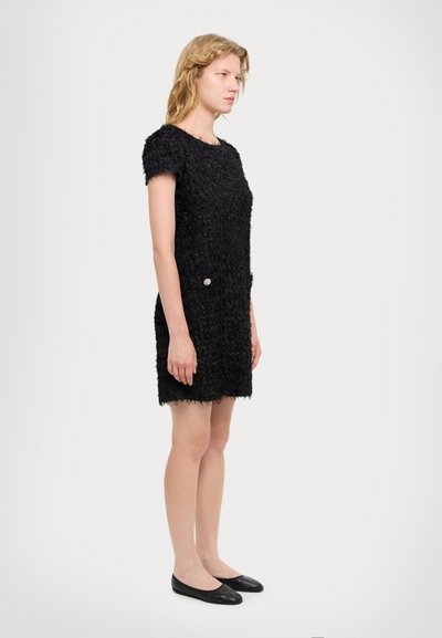 Vestido negro texturizado con mangas cortas, que presenta una tela suave y un detalle de botón decorativo en la cintura. Bailarinas negras completan el look.